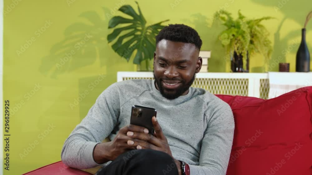 Joyful smiling african man browsing fun content on smartphone staying lazy at home. Portrait of handsome millennial guy using modern iphone in stylish living room.