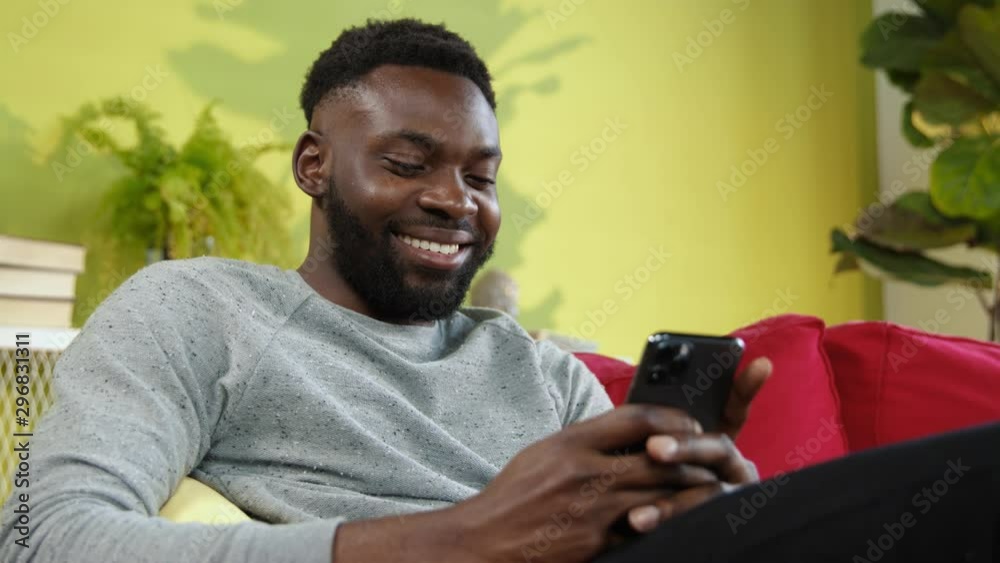 Pleased smiling afro-american man using smartphone chatting with friends laughing enjoying afternoon time at home.