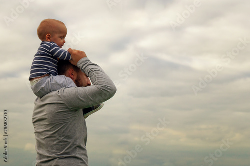 A child on his father’s neck. Walk near the water. Baby and dad against the sky.