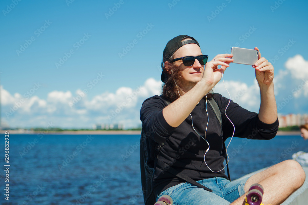 A young man sitting on the embankment with a yellow skateboard on his lap makes a photo on smartphone of the cityscape.