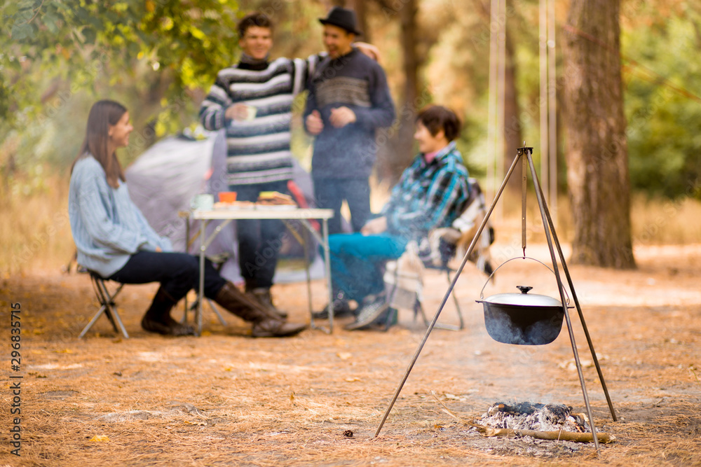Group of people sitting at tourist table and eating food cooked on fire. Camp food. Group of young people in casual wear resting in autumn forest Group of people sitting at tourist table and eating food cooked on fire. Camp food. Group of young people in casual wear resting in autumn forest