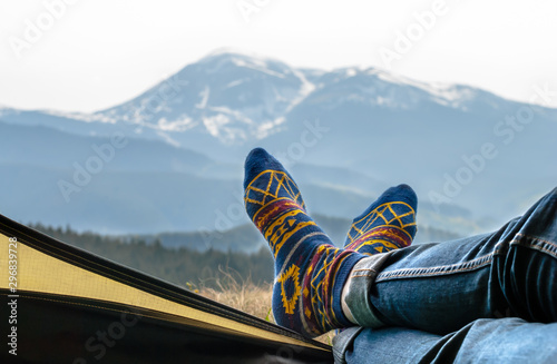Women's legs in blue jeans and fun socks with a bright yellow pattern lie on the edge of the yellow tent. Mountain ranges covered with green forest and snow. View from the open tent.