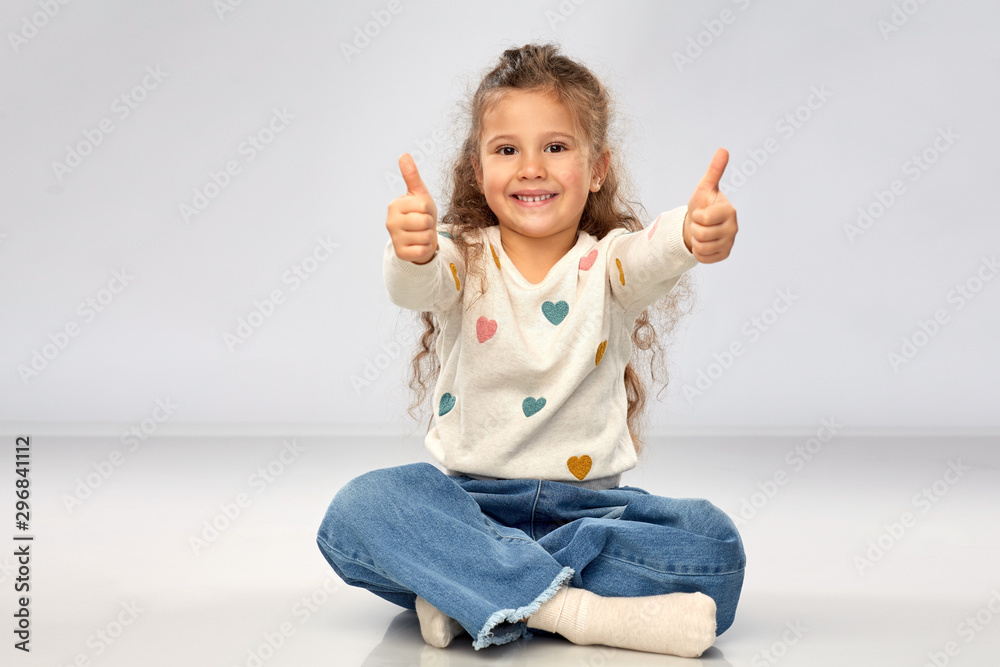 gesture, childhood and people concept - beautiful smiling girl sitting on floor and showing thumbs up over grey background gesture, childhood and people concept - beautiful smiling girl sitting on floor and showing thumbs up over grey background