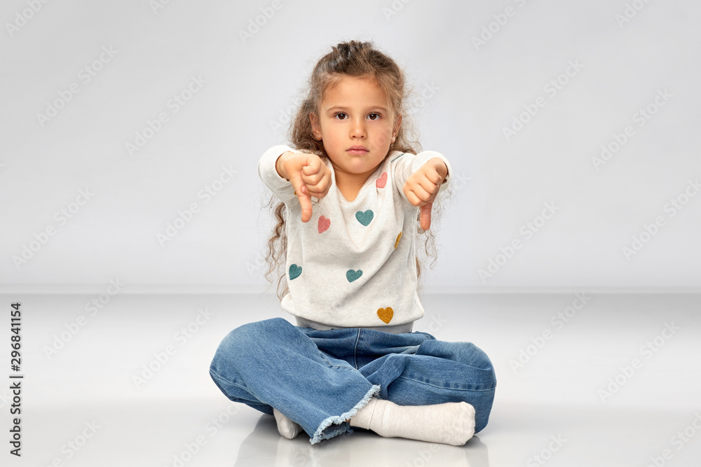 gesture, childhood and people concept - sad girl sitting on floor and showing thumbs down over grey background gesture, childhood and people concept - sad girl sitting on floor and showing thumbs down over grey background