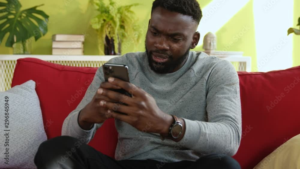 Afro-american man using modern smartphone resting on red sofa in the house. Excited handsome guy receiving great news cheering up and celebrating business success. Afro-american man using modern smartphone resting on red sofa in the house. Excited handsome guy receiving great news cheering up and celebrating business success.