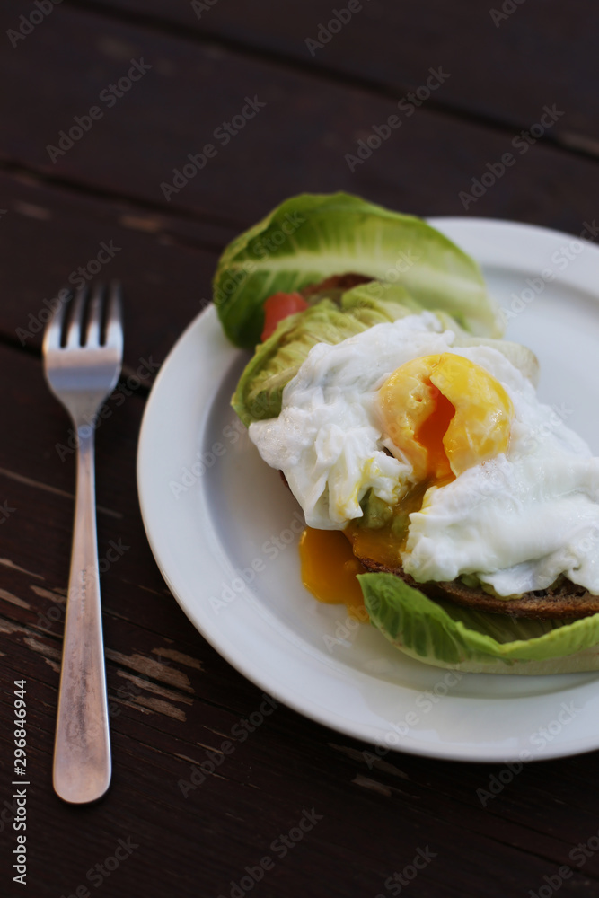 Close-up flat lay of delicious poached egg with liquid yolk on toast with fresh salad leaves and avocado at dark rustic brown wooden background/ table. Top view of healthy protein breakfast or lunch. Close-up flat lay of delicious poached egg with liquid yolk on toast with fresh salad leaves and avocado at dark rustic brown wooden background/ table. Top view of healthy protein breakfast or lunch.