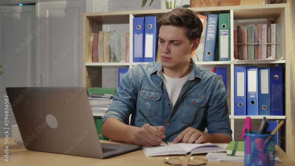 Serious smart young man business specialist working on notebook making notes managing financial deals in cozy company office. Serious smart young man business specialist working on notebook making notes managing financial deals in cozy company office.