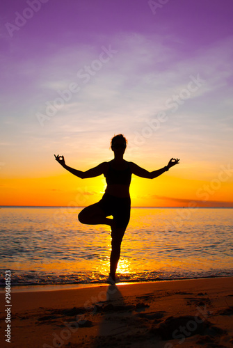 Fototapeta Naklejka Na Ścianę i Meble -  A young lady practices yoga on the beach in front of the setting sun. The Caribbean sea is tranquil as it laps the sandy coastline of the idyllic island