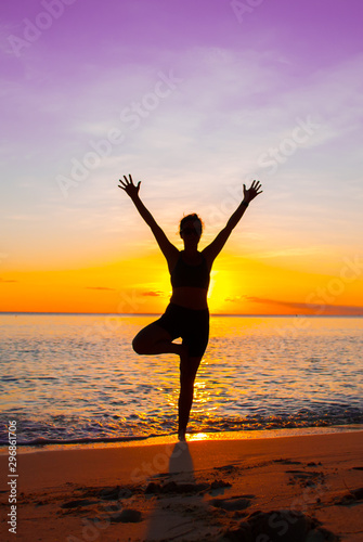 A young lady practices yoga on the beach in front of the setting sun. The Caribbean sea is tranquil as it laps the sandy coastline of the idyllic island
