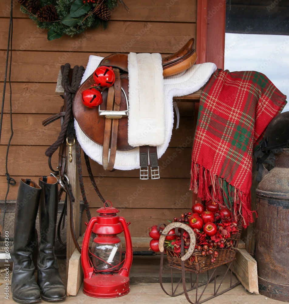 Obraz premium Portrait of a Black Great Dane resting on a farmhouse porch beside a saddle rack with English tack including saddle, bridle, riding boots decorated for Christmas- bells, lantern and plaid blanket