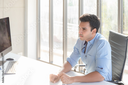 A handsome Asian guy wearing a blue shirt with a serious face, sitting at a desk with a computer placed on the desk in the office working room in the morning.