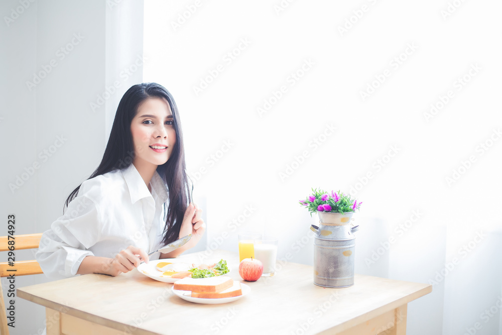A beautiful Asian woman, a tan, wearing a long-sleeved white shirt and shorts, and on her hand holding a fork and a knife, is eating breakfast in a bedroom at a hotel in Thailand