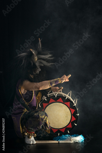 Demon from Japanese mythology. Full lenght portrait of an artist drummer Taiko in a wig with horns and make-up sits on stage and shakes head against a dark background.