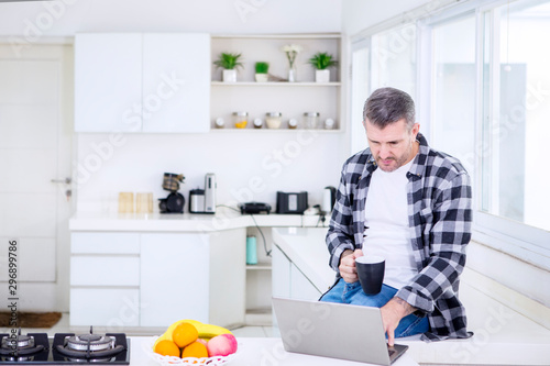 Man holding coffee and using a laptop in kitchen