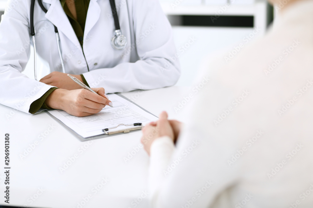 Woman doctor and patient sitting and talking at medical examination at hospital office, close-up. Therapist filling up medication history records. Medicine and healthcare concept Woman doctor and patient sitting and talking at medical examination at hospital office, close-up. Therapist filling up medication history records. Medicine and healthcare concept