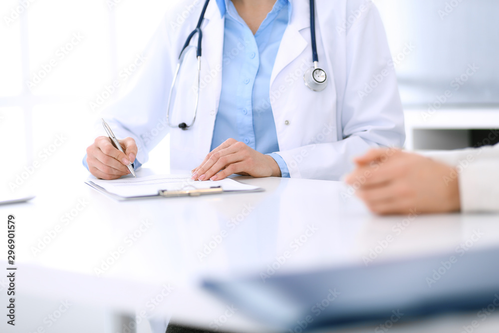 Woman doctor and patient sitting and talking at medical examination at hospital office, close-up. Physician filling up medication history records. Medicine and healthcare concept Woman doctor and patient sitting and talking at medical examination at hospital office, close-up. Physician filling up medication history records. Medicine and healthcare concept