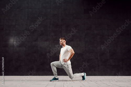 Side view of fit handsome muscular bearded focused sportsman in tracksuit doing lunges outdoors.