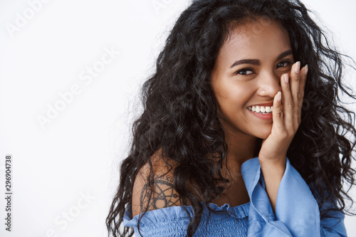 Tenderness, beauty and wellbeing concept. Charming feminine african american woman with black curly hairstyle, touching cheek, blushing happily, look camera giggling, stand white background