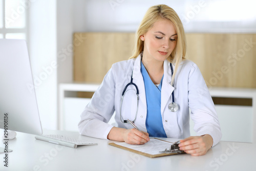 Doctor woman at work while sitting at the desk in hospital or clinic. Blonde cheerful physician filling up medical records form. Data and best service in medicine and healthcare