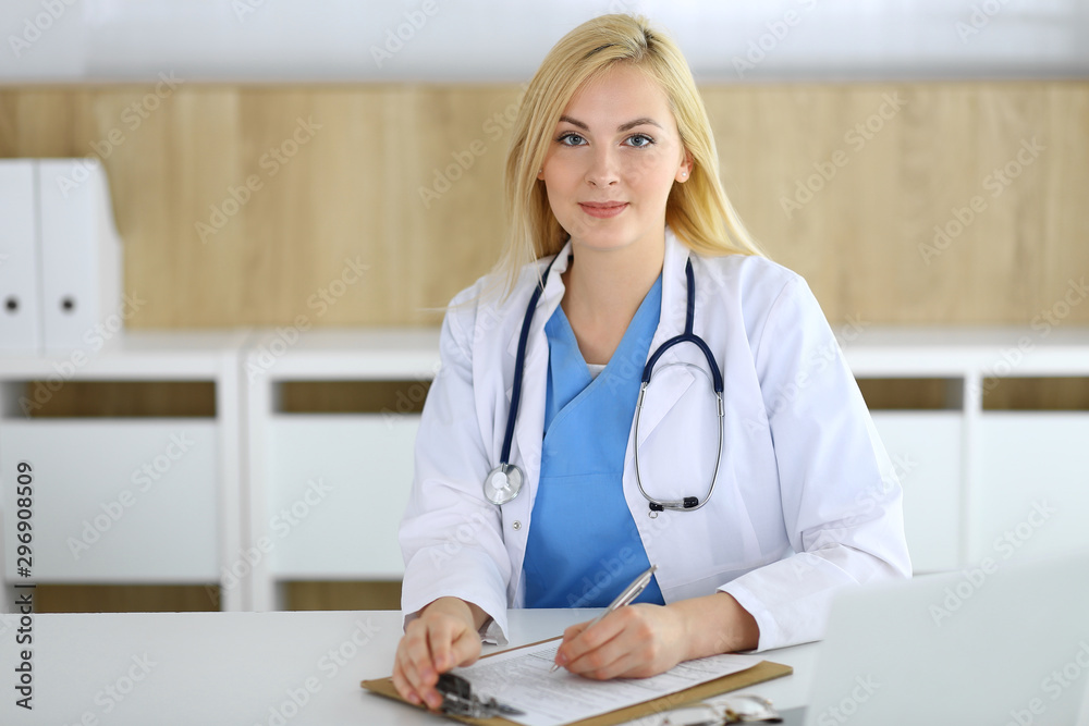 Doctor woman at work while sitting at the desk in hospital or clinic. Blonde cheerful physician filling up medical records form. Data and best service in medicine and healthcare Doctor woman at work while sitting at the desk in hospital or clinic. Blonde cheerful physician filling up medical records form. Data and best service in medicine and healthcare