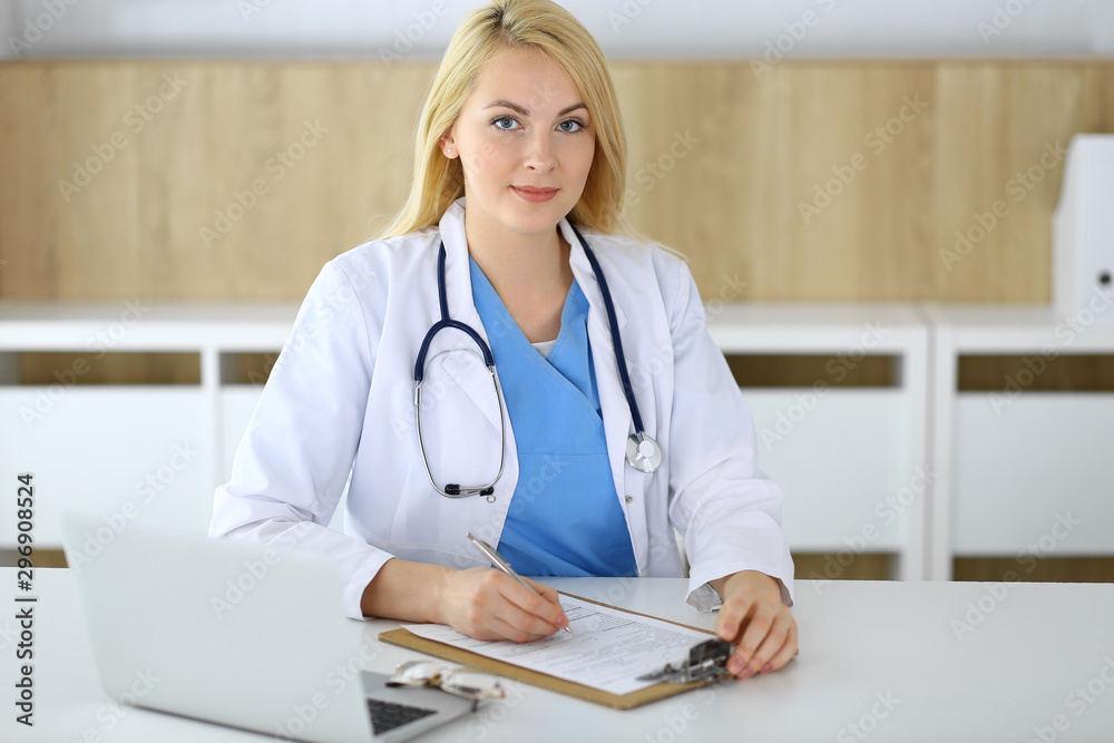 Doctor woman at work while sitting at the desk in hospital or clinic. Blonde cheerful physician filling up medical records form. Data and best service in medicine and healthcare Doctor woman at work while sitting at the desk in hospital or clinic. Blonde cheerful physician filling up medical records form. Data and best service in medicine and healthcare