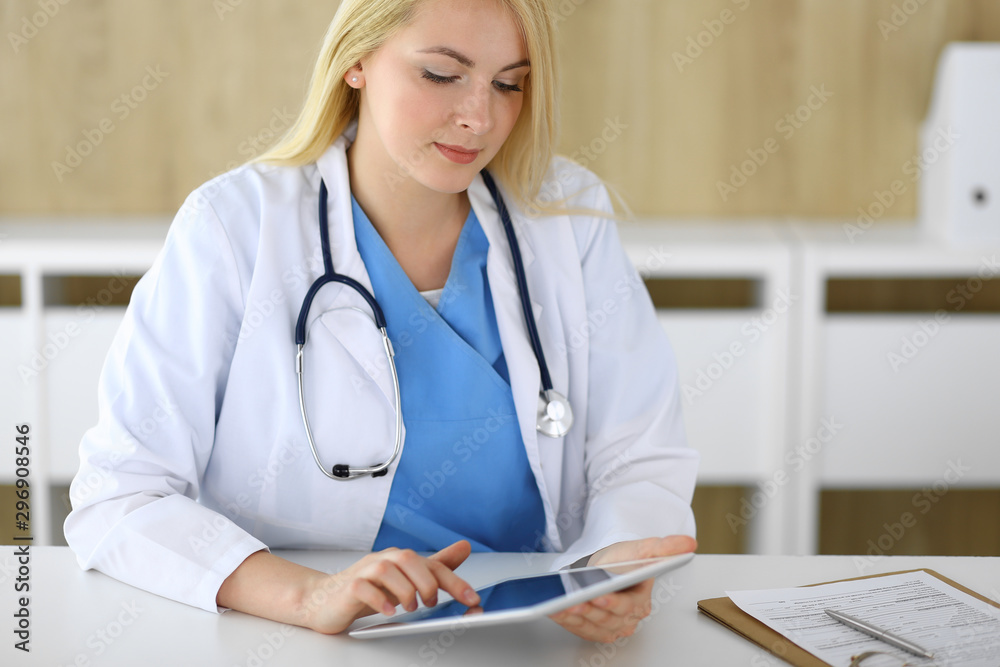 Woman doctor using tablet pc in hospital or clinic. Blonde therapist cheerfully smiling. Data in medicine and healthcare concept Woman doctor using tablet pc in hospital or clinic. Blonde therapist cheerfully smiling. Data in medicine and healthcare concept