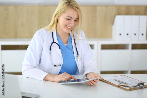 Woman doctor using tablet pc in hospital or clinic. Blonde therapist cheerfully smiling. Data in medicine and healthcare concept