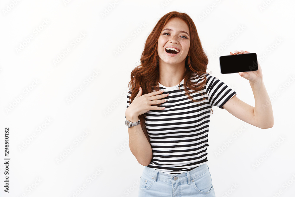 Grateful and flattered cute redhead girl in striped t-shirt, showing smartphone display horizontally, touching chest thankful and touched, smiling happily, laughing and gazing camera Grateful and flattered cute redhead girl in striped t-shirt, showing smartphone display horizontally, touching chest thankful and touched, smiling happily, laughing and gazing camera