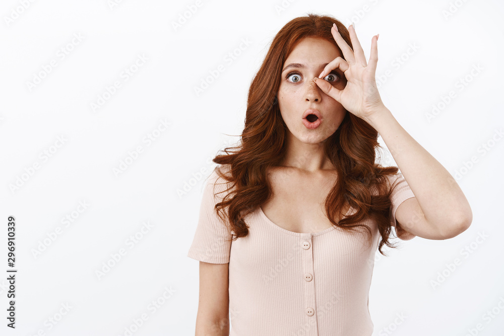 Fototapeta premium Fascinated cute ginger girl in blouse with freckles, stare impressed, looking through fingers surprised and speechless, checking our breathtaking cool thing, standing white background amused