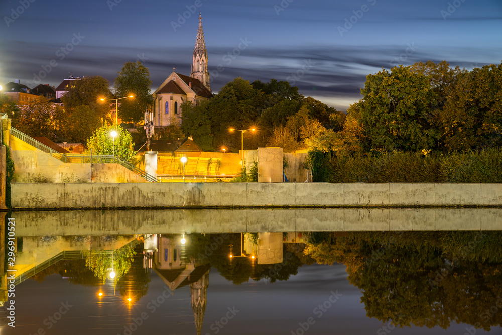 Germany, Beautiful berger church reflecting in silent glassy neckar river water by night in stuttgart bad canstatt surrounded by green trees illuminated by street lamps
