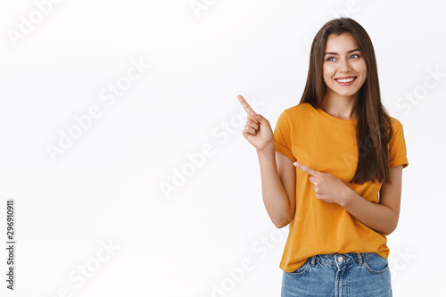 Cheerful, carefree happy young friendly woman in yellow t-shirt, smiling laughing joyfully as looking enthusiastic upper left corner, pointing with fingers at someone or promo, white background