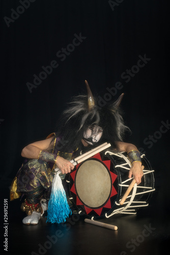 Taiko drummer in a wig and a demon mask on stage squarts with drumstick and drum and listen on a black background. Demon from Japanese mythology.