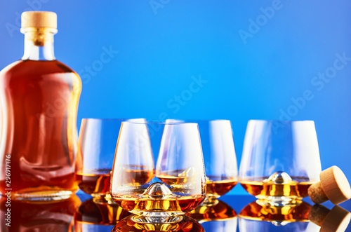 Crystal glass of whiskey on the glass surface and blue background. In the background  three glasses of whiskey  a full bottle and a wooden cork. The composition is reflected on the surface.