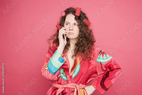 Image of angry millennial housewife clenches fists, looks with irritation aside, annoyed by noisy people who disturb her, wants to rest in peaceful atmosphere, isolated with curlers on head on pink.