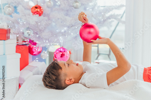 Up-view shot of little child laying under christmas tree, touching balls, looking at his reflection in ornaments glass.