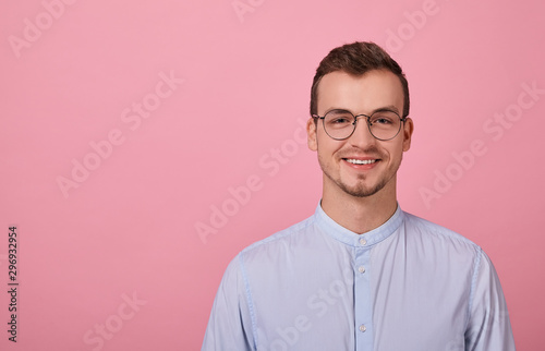 A decent young man in a sky blue shirt and computer glasses stands on a pink background. Student, employee of the year.