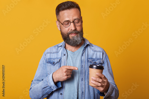 Photo of attractive man pointing on cup of coffee or tea, looking at disponsable mug, posing isolated over yellow background, handsome male with coffee cup dresses jacket. People lifestyle concept. photo