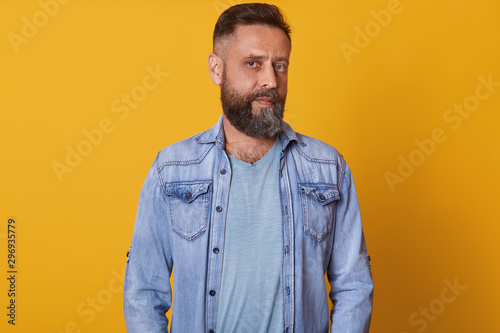 Horizontal shot of handsome young bearded man looking at camera while standing against yellow studio background, attractive male turning his head aside, wearing stylish clothing. People concept.