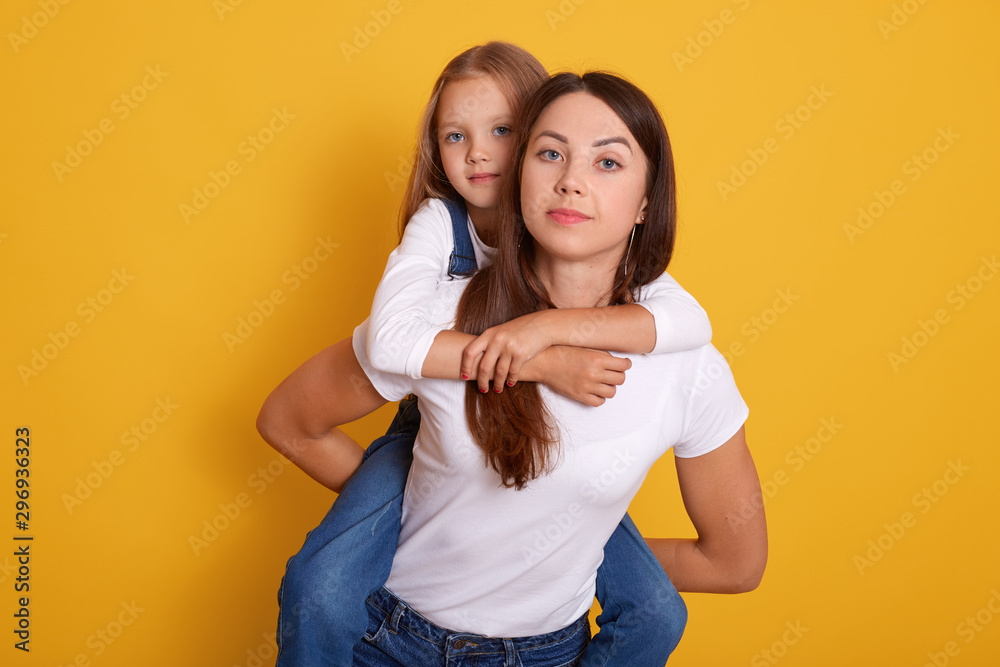 Close up portrait of mother giving piggyback ride to her cute daughter and being in good mood, models posing isolated over yellow background, woamn wearing white t shirt holding her blonde kid. Close up portrait of mother giving piggyback ride to her cute daughter and being in good mood, models posing isolated over yellow background, woamn wearing white t shirt holding her blonde kid.