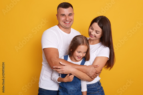 Close up portrait of young Caucasian family.Father, mother and charming daughter. Parents hugging little cute child dressed denim overalls. Woman and man smiling, expressing love and happyness.