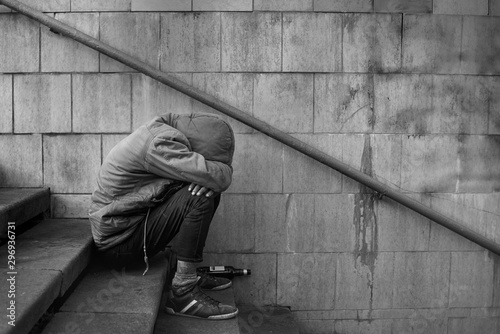 Drunk homeless man covered his face with his hands and sits on the stairs in the underpass, black and white photo.