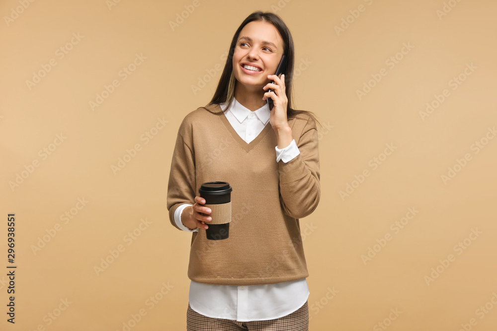 Attractive happy girl smiling and holding coffee cup while talking on phone, isolated on brown background. Conversation concept. Attractive happy girl smiling and holding coffee cup while talking on phone, isolated on brown background. Conversation concept.