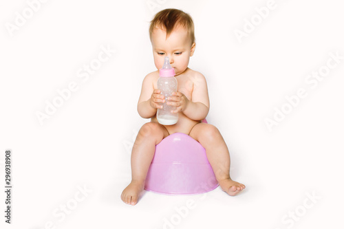 Charming photo of cute baby girl holding a boottle with baby milk and sitting on a pink potty. Isolated on white background