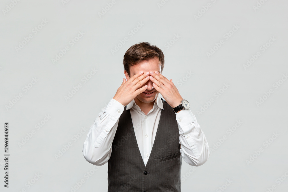 Handsome man in a white shirt and grey vest hiding behind his hands isolated over grey background. Handsome man in a white shirt and grey vest hiding behind his hands isolated over grey background.