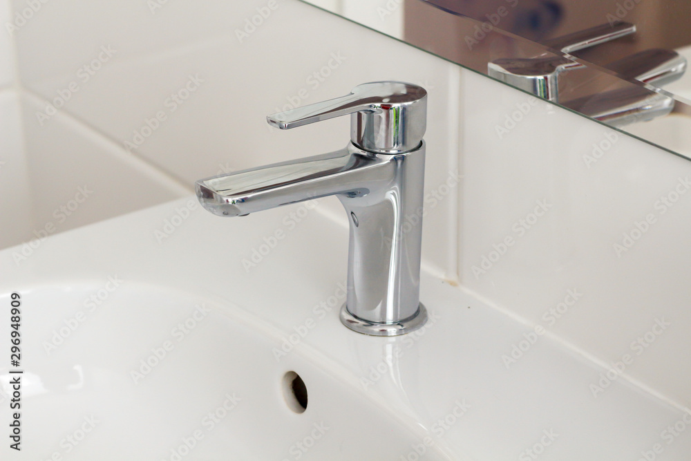 Silver metal shiny faucet water switch and ceramic sink with big mirror on background in hotel home spa bathroom with white tiles on wall. Bathroom luxury interior. Soft selective blurred focus Silver metal shiny faucet water switch and ceramic sink with big mirror on background in hotel home spa bathroom with white tiles on wall. Bathroom luxury interior. Soft selective blurred focus