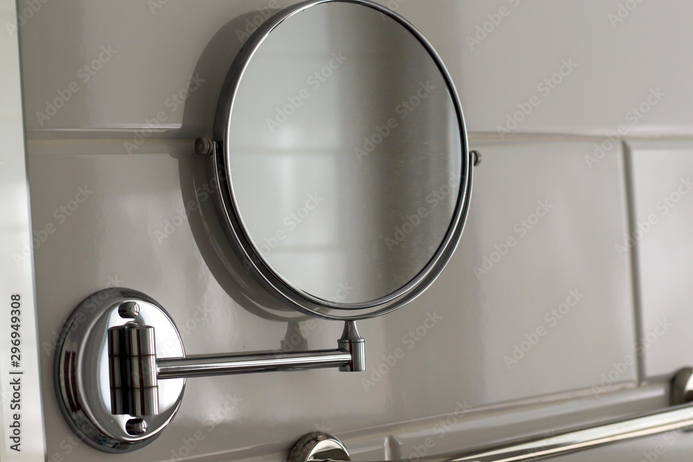 Round magnifying circle mirror hanging on the wall in the bathroom of the hotel spa house on white tiles. Bathroom luxury interior. Round magnifying circle mirror hanging on the wall in the bathroom of the hotel spa house on white tiles. Bathroom luxury interior.