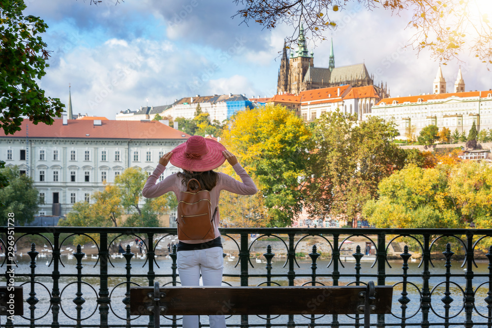 Eine Touristin mit rotem Sonnenhut steht am Ifer der Moldau und schaut auf das Schloss von Prag an einem sonnigen Herbsttag, Tschechien