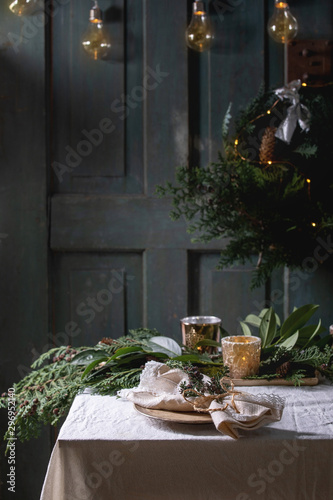 Christmas or New year table setting with empty ceramic plates, napkins, Christmas thuja wreath, luminous garland and burning candles on white tablecloth. Holiday mood