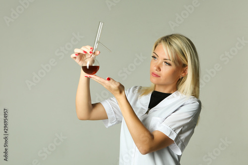 beautiful female lab technician holding a flask with a chemical reagent on gray background with copy space. beautiful blonde in a white coat looks at a chemical solution in a bottle