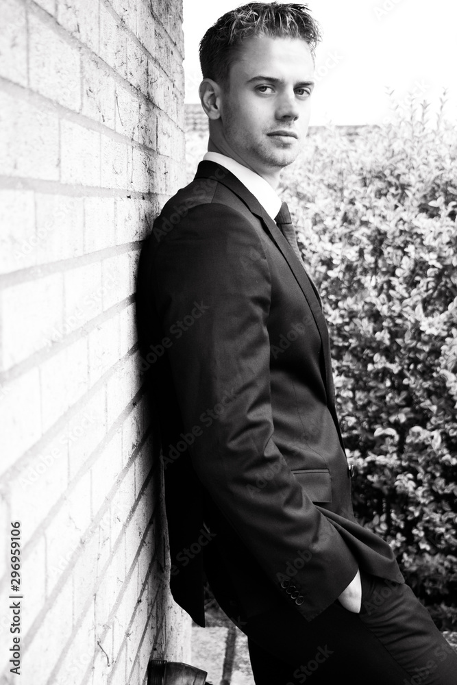 Black and white portrait of handsome man wearing suit standing next to brick wall looking at camera